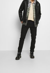 Giacca di pelle nera sopra una camicia a quadri grigia, una maglietta beige e jeans neri. Completano il look stivaletti neri con suola spessa.