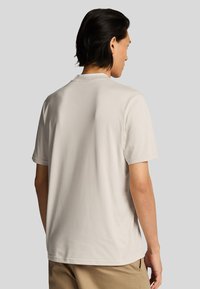 Kurzärmliges, hellbeiges T-Shirt aus weicher Baumwolle, mit Rundhalsausschnitt und lässiger Passform, von hinten vor einem grauen Hintergrund gezeigt.