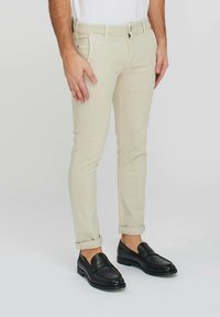 Jeans slim fit beige chiaro con una texture liscia, dotati di chiusura con bottone e orli piegati, abbinati a mocassini neri.
