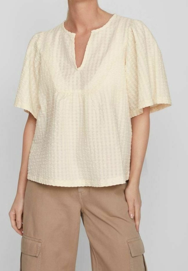 Krämfärgad kortärmad topp med V-hals, texturerad tyg och lös passform. Har en samlad söm under bystlinjen.