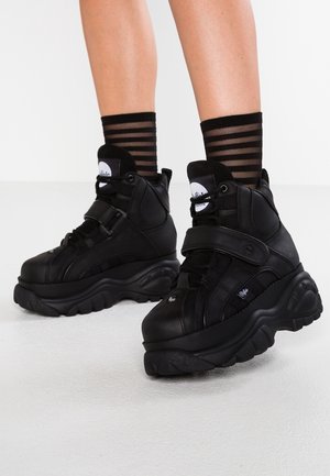 Personne portant des baskets plateformes noires épaisses avec un maintien élevé de la cheville et des chaussettes noires transparentes à rayures, debout sur un sol clair.