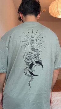 T-shirt verde menta con grafica di un serpente nero e design di luna crescente e raggi di sole sul retro. Maniche corte e vestibilità comoda.