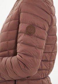 Brun pufferjakke med quiltet design og et rundt læderlogo-patch på venstre arm, der viser en glat, skinnende tekstur.
