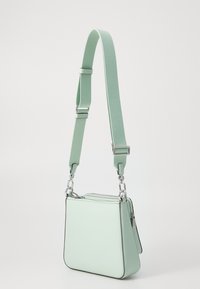 Borsa quadrata verde menta con tracolla texturizzata, dotata di una cerniera e bordi eleganti, posizionata su uno sfondo uniforme.