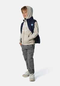Chaleco acolchado azul marino sobre una sudadera con capucha beige, acompañado de pantalones cargo grises y zapatillas claras. Cuenta con un parche de logo en el chaleco.