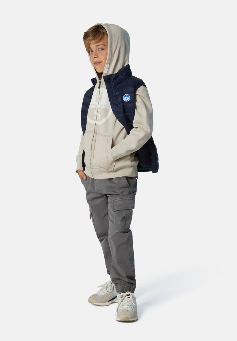 Chaleco acolchado azul marino sobre una sudadera con capucha beige, acompañado de pantalones cargo grises y zapatillas claras. Cuenta con un parche de logo en el chaleco.
