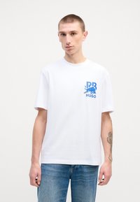 T-shirt bianco in cotone con un design grafico blu sul lato sinistro del petto con "PR" e "HUGO". Maniche corte e vestibilità rilassata.