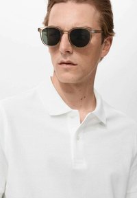 Sonnenbrille mit runden schwarzen Gläsern und klaren geometrischen Rahmen. Weißes Poloshirt mit einer strukturierten, gewebten Musterung und einer Drei-Knopf-Leiste.