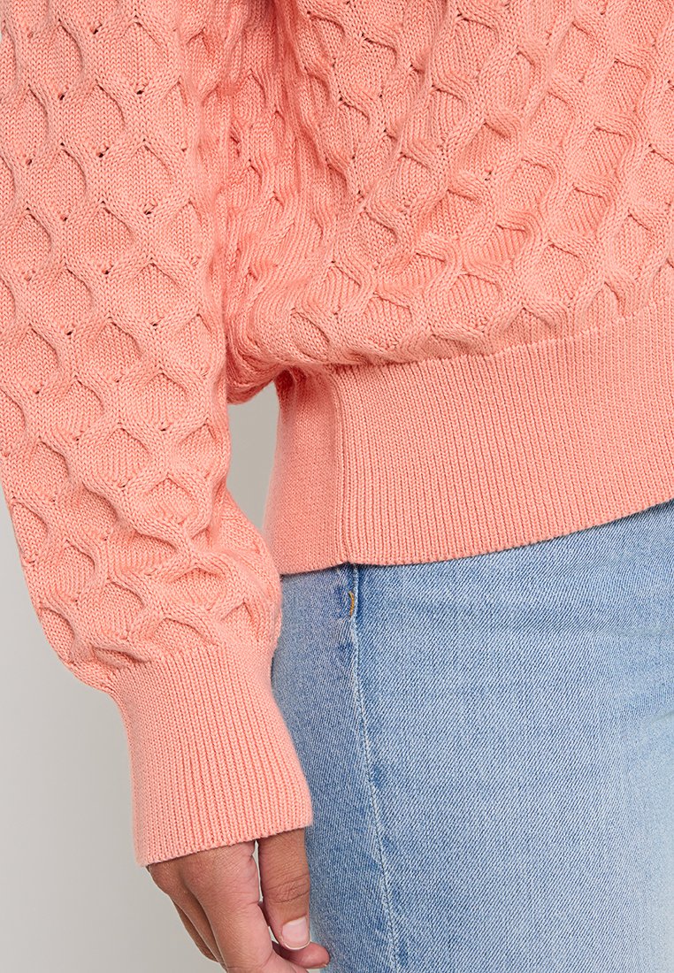 Teksturowany, koralowy sweter dzianinowy z diamentowym wzorem, ściągaczami przy rękawach i dopasowanym pasem; w połączeniu z jasnoniebieskimi dżinsami.
