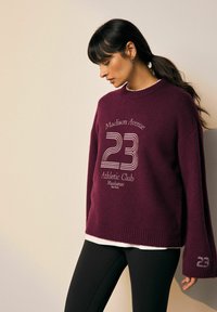 Bordo crewneck svetar s grafičkim tekstom "Madison Avenue 23 Athletic Club" i detaljnim šavovima. Nositi preko bijele majice i crnih tajica.