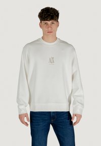 Weißes Sweatshirt aus weichem Stoff mit Rundhalsausschnitt, mit einem kleinen goldenen Logo auf der Brust, kombiniert mit dunkelblauen Jeans.