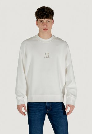 Sudadera blanca de tejido suave con cuello redondo, que presenta un pequeño logo dorado en el pecho, combinada con jeans de color azul oscuro.