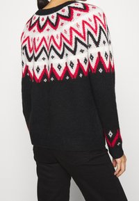 Gemusterter Strickpullover mit einem weißen, roten und schwarzen geometrischen Design im oberen Bereich, das unten in ein einheitliches Schwarz übergeht.