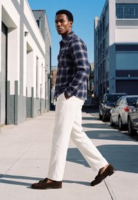 Homme marchant sur un trottoir urbain portant une chemise à carreaux bleue, un pantalon blanc et des mocassins marron, les mains dans les poches.