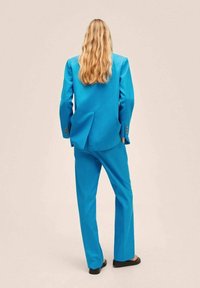 Costume bleu vif avec un blazer ajusté et un pantalon assorti, présentant une fente unique dans le dos, conçu dans un tissu lisse.