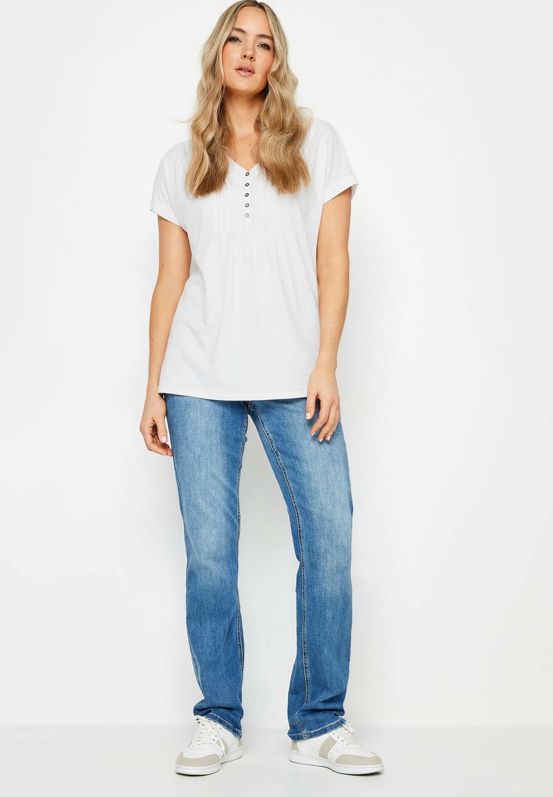 Top bianco a maniche corte con scollo a V e dettagli con bottoni, abbinato a jeans in denim azzurro chiaro a gamba dritta e sneakers bianche.