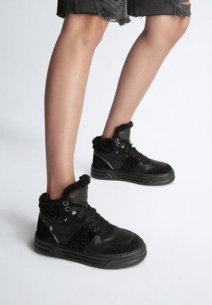 Jambes portant des baskets montantes noires avec une doublure en fourrure et des shorts en jean noirs déchirés sur un fond blanc uni.