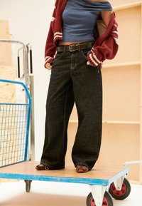 Person wearing wide-leg black jeans, blue off-shoulder top, and red cardigan stands on a wooden platform cart with red wheels.
