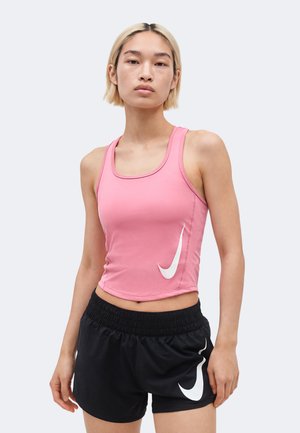 Jeune personne aux cheveux blonds courts portant un haut rose sans manches et un short de sport noir avec des logos blancs, debout devant un fond uni.