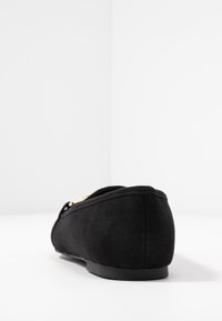 Chaussure plate en daim noir avec un petit talon, vue de dos sur une surface blanche contre un fond blanc.