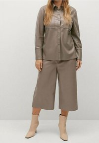 Hnědá košile z umělé kůže s knoflíkovým zapínáním a dvěma náprsními kapsami, v kombinaci s odpovídajícími culottes a béžovými kotníkovými botami. Hladká textura.