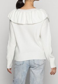 Maglione bianco a coste con un ampio colletto ruffle e maniche lunghe, abbinato a jeans in denim blu chiaro. Texture morbida e vestibilità rilassata.