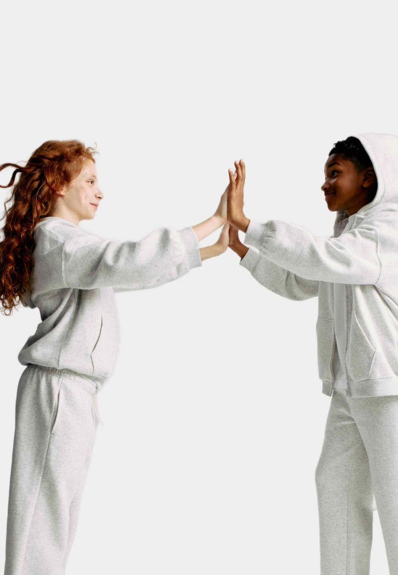 Due bambini in felpe e pantaloni tuta grigio chiaro, dal tessuto morbido, si affrontano, toccandosi i palmi delle mani, con capelli ricci rossi e capelli corti scuri.