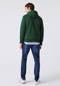 Hombre con sudadera con capucha verde oscuro, jeans azules y zapatillas blancas, de pie de espaldas contra un fondo simple.