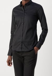 Camicia nera con bottone, maniche lunghe, texture liscia, colletto classico, design aderente, piccoli bottoni, abbinata a pantaloni neri.