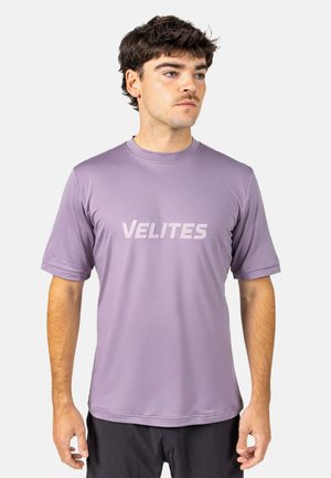 Hombre mirando hacia la derecha, vistiendo una camisa de manga corta lavanda con "VELITES" impreso en letras mayúsculas en el pecho.