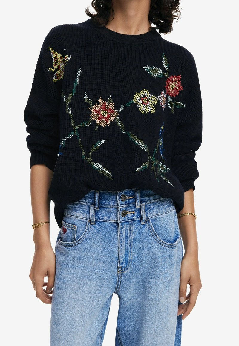 Personne portant un pull en tricot noir avec des motifs floraux colorés brodés en point de croix et un jean taille haute bleu clair à double bouton.