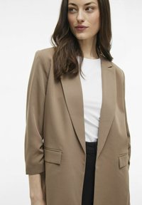 Blazer marrón de corte ajustado, con frontales abiertos y solapas con muesca, que cuenta con dos bolsillos frontales y una tela suave y texturizada.