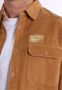 Chemise en velours côtelé marron avec une surface texturée, boutonnage sur le devant, poche poitrine unique et une étiquette cousue avec la marque "OXBOW".