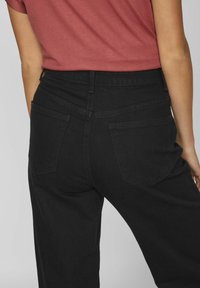 Črne denim hlače z visokim pasom, ki imajo dve zadnji žepki in subtilne šivane detajle. Tkanina ima gladko, mat teksturo.
