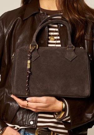 Woman holding dark brown suede handbag with braided charm, wearing brown leather jacket, striped shirt, gold jewelry, and rings.