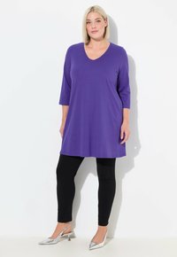 Lila Tunika mit V-Ausschnitt und drei Viertelärmeln, kombiniert mit schwarzen Leggings und silbernen, metallic-finish High Heels. Glatte Stoffoberfläche.