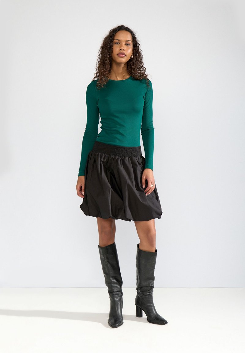 Vrouw met krullend haar, gekleed in een teal top met lange mouwen, een zwarte geplooide rok en zwarte kniehoge laarzen met hak, staand tegen een witte achtergrond.