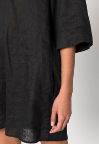 Bras et main d'une personne portant une robe ample en lin noir avec des manches mi-longues sur un fond uni.