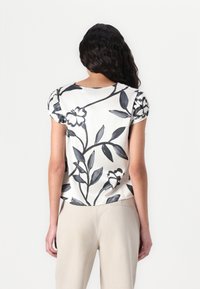 Blusa a maniche corte di colore bianco con un motivo floreale nero. Presenta maniche arricciate e un collo rotondo, realizzata in un tessuto morbido e liscio.