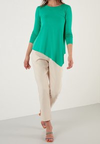 Turkosh långärmad topp med asymmetrisk nederkant, som kombineras med beige byxor och orange hälskor med remmar. Slät tyg med en figurnära silhuett.