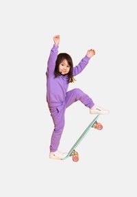 Fioletowa bluza i spodnie. Dziecko balansuje na turkusowej deskorolce z różowymi kółkami. Białe trampki. Neutralne tło.