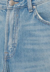 Blugi din denim albastru deschis văzuți de aproape, prezentând nasturele, bucla pentru curea, cusăturile portocalii și buzunarul mic frontal cu un ușor detaliu de uzură a țesăturii.