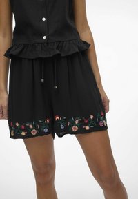 Combinaison noire avec ourlet à volants, ornée de broderies florales colorées en bas. Fabriquée en tissu léger avec une taille à cordon.