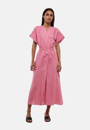 Femme debout portant une robe midi rose à manches courtes avec une taille cintrée et une fente frontale, associée à des talons noirs ouverts à l'avant.