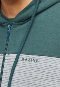 Osoba ubrana w zieloną i jasnoszaro paskowaną bluzę z kapturem na zamek, z haftem "MAZINE" na piersi.