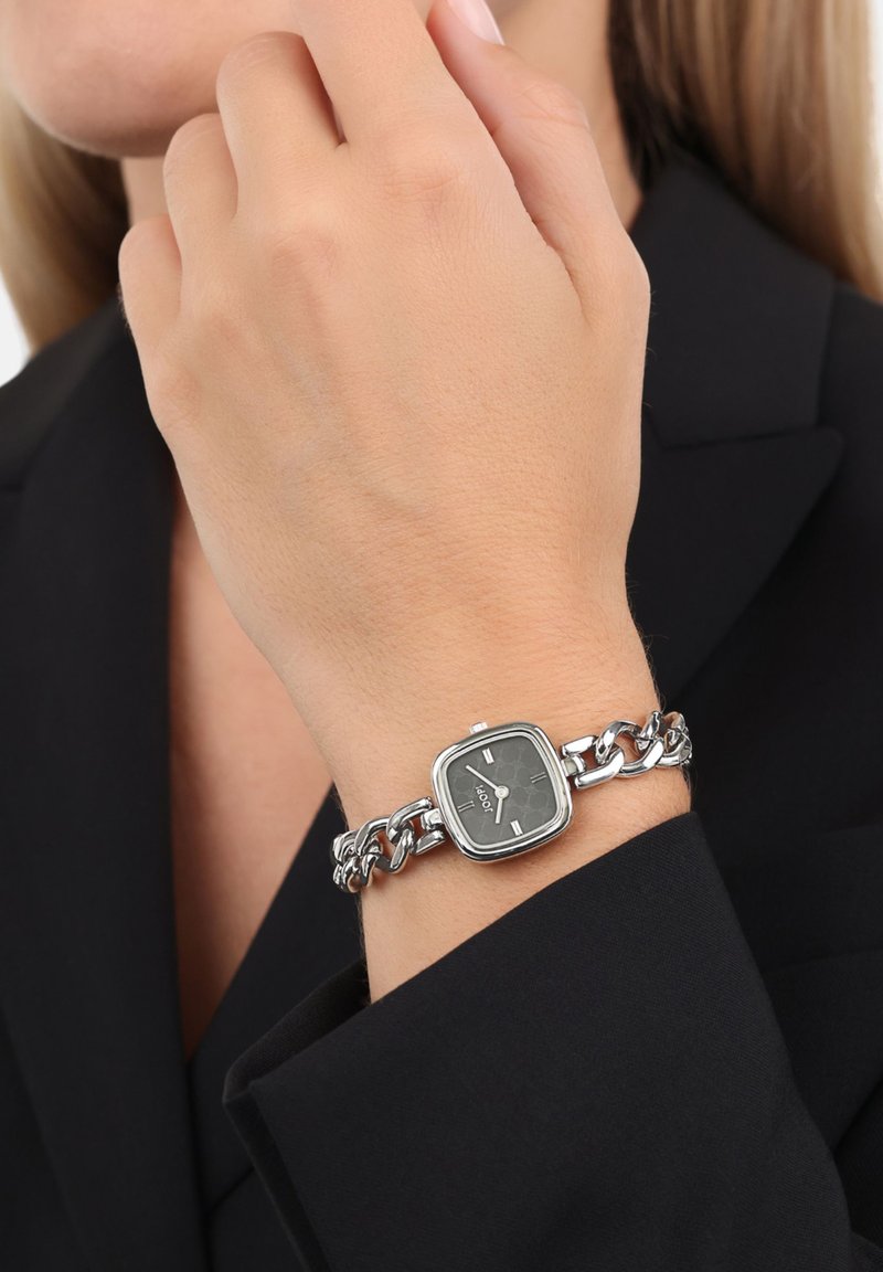 Silberne Metallarmbanduhr mit quadratischem schwarzem Zifferblatt, Kettenarmband und weißen Stundenmarkierungen, getragen an einer hellen Hand gegen ein dunkles Hemd.