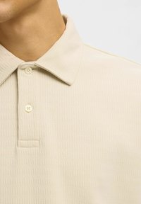 Kadar u blizini bež teksturiranog polo majice s dva kremasta gumba i ovratnikom, prikazujući dio vrata i ramena.