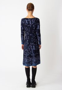 Robe marine à manches longues, motifs floraux dans des bleus plus clairs, jupe plissée et taille cintrée. Portée avec des chaussettes noires jusqu'aux genoux et des chaussures épaisses.