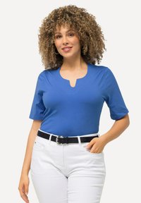 Modrý top s krátkým rukávem a výstřihem do V, vyrobený z hladkého materiálu. V kombinaci s bílými kalhotami a černým páskem se stříbrnou přezkou.