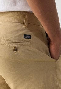 Khaki püksid, millel on tekstureeritud kangas, ühe tagatasku nupp, ning must silt, millel on kirjas "ÜKSASTANDE ÜKSAASTA".